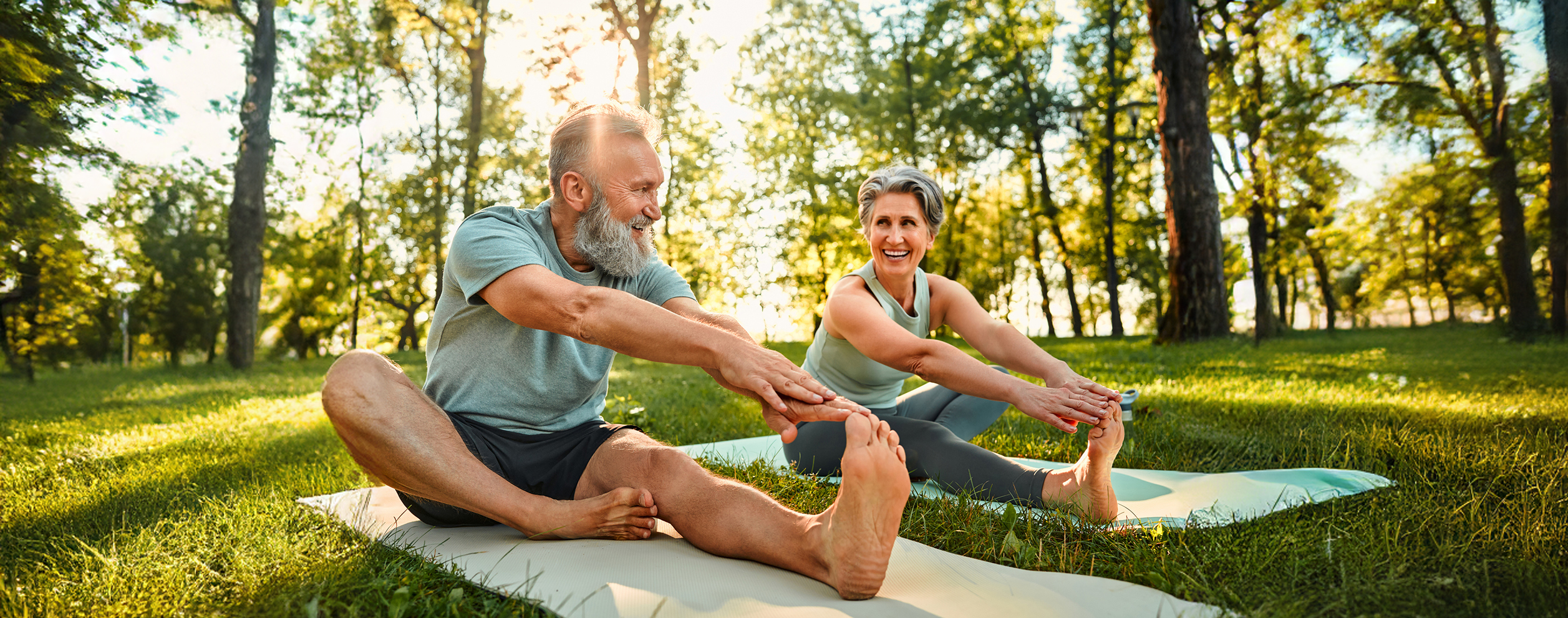 Sporty man and woman with grey hair stretching on yoga mats with hands to one leg during outdoors workout.